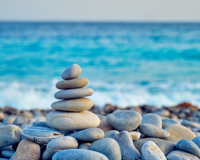 A serene image of smooth pebbles stacked in balance on a sandy beach, symbolizing harmony, stability, and a whole-body approach to wellness. The peaceful coastal backdrop reflects the connection between nature, health, and holistic well-being.